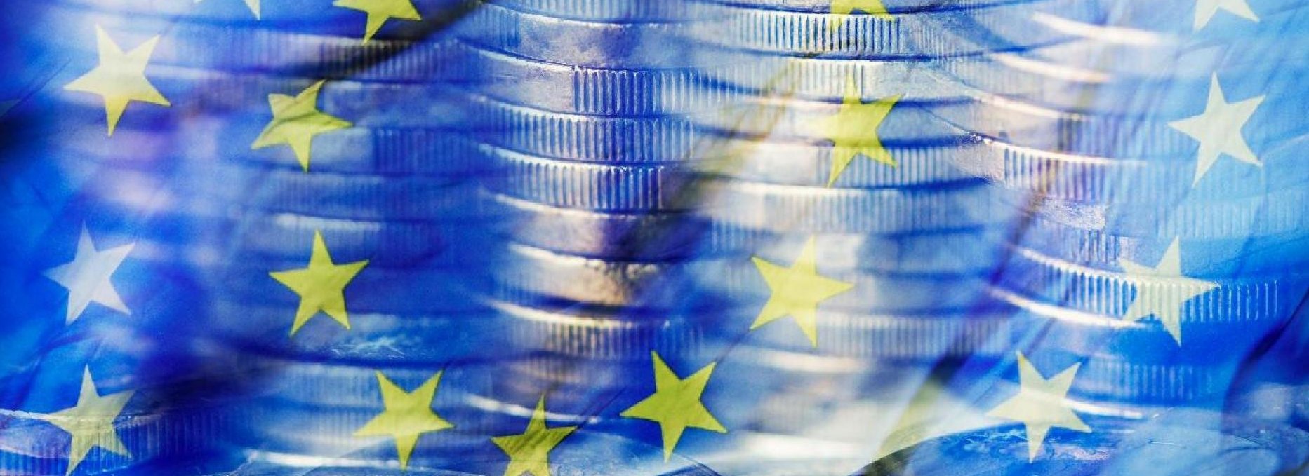 Zu sehen sind gestapelte ein Euro-Münzen, die hinter einer transparenten EU-Flagge sind.