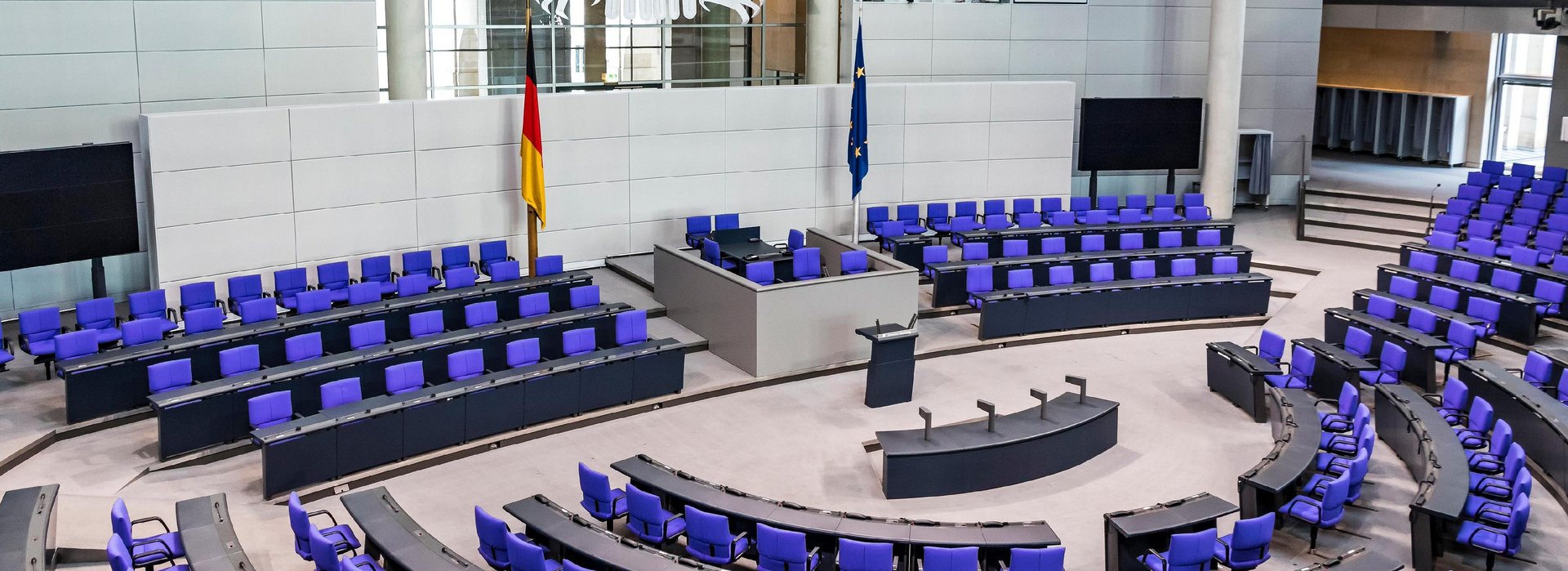 A photograph of the empty plenary hall of the German Bundestag. You can see the blue seating for the parliamentary groups as well as the seats for the government and the parliamentary presidents.
