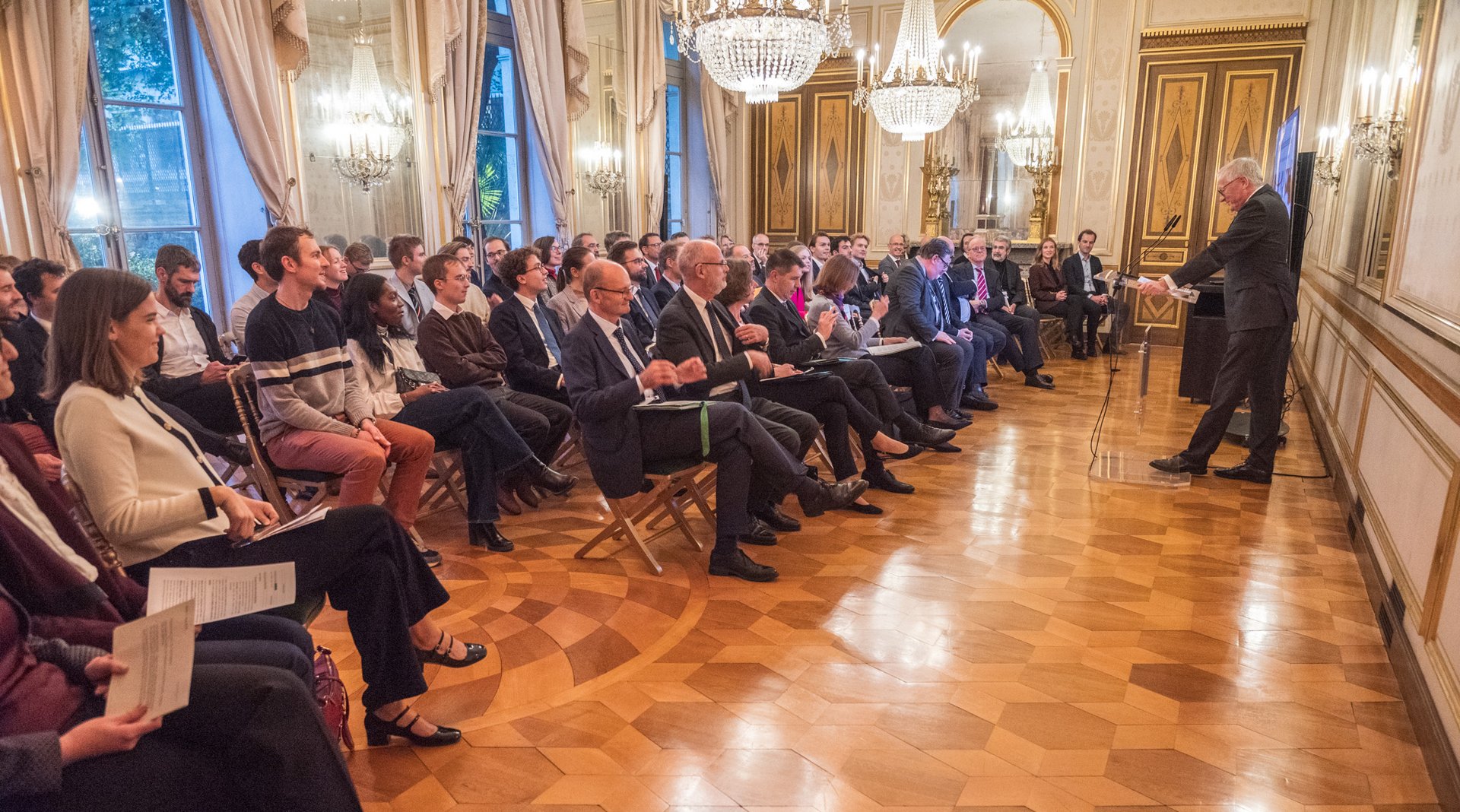Zu sehen ist ein prunkvoller Saal im "Empire-Stil" des franz&ouml;sischen 18. Jahrhunderts. Ein Mann im Anzug h&auml;lt vor einer auf Stuhlreihen sitzenden Gruppe einen Vortrag. 