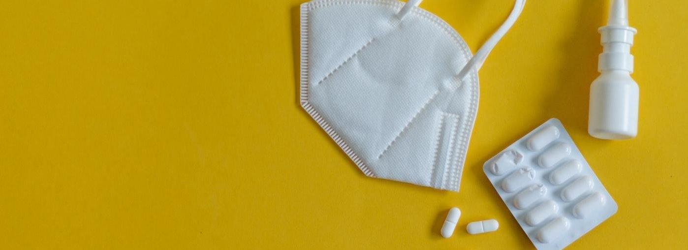 On a yellow background are a white FFP2 mask, a white nasal spray, a glass clinical thermometer and a tablet blister, three tablets of which are already lying individually next to the blister.